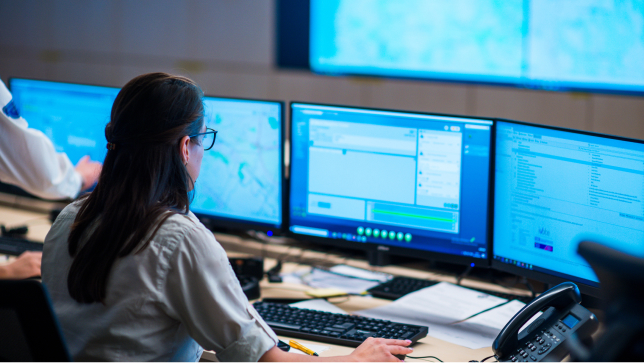 Una mujer en un centro de comando viendo varias pantallas de ordenador y moviendo el ratón de color negro 