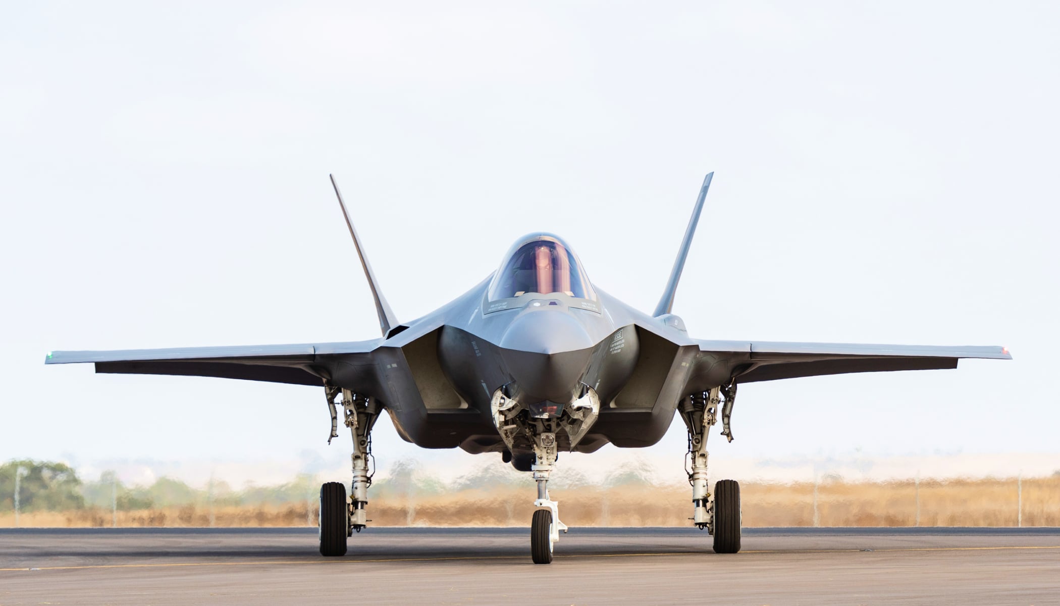 Fotografía de un moderno avión de combate gris estacionado en una pista listo para volar, tomada de frente con el morro de la nave apuntando hacia el espectador. Al fondo se ve un campo llano de color marrón bajo un cielo claro
