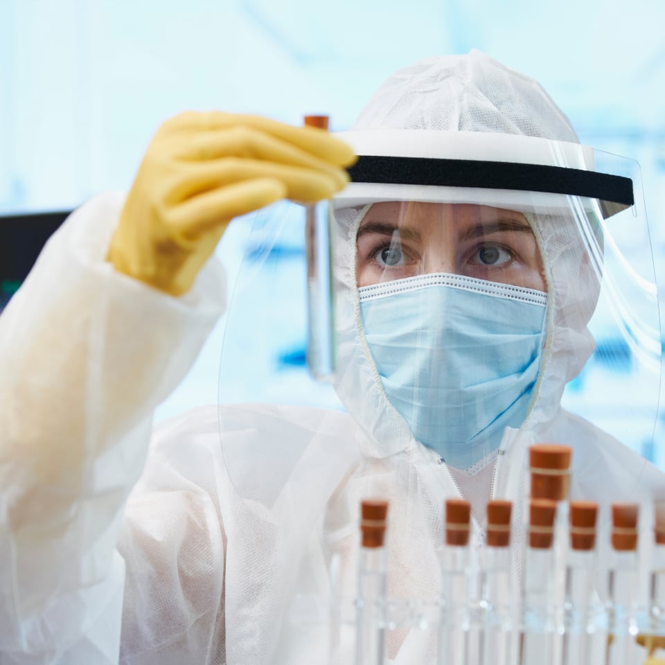A researcher wearing a white full-body containment suit and yellow gloves holds a test tube up to the light