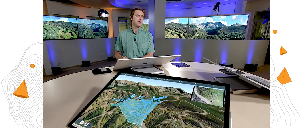 Hombre de pie en una gran mesa redonda delante de un monitor de ordenador grande con 2 grandes monitores de televisión que muestra una imagen digital en 3D