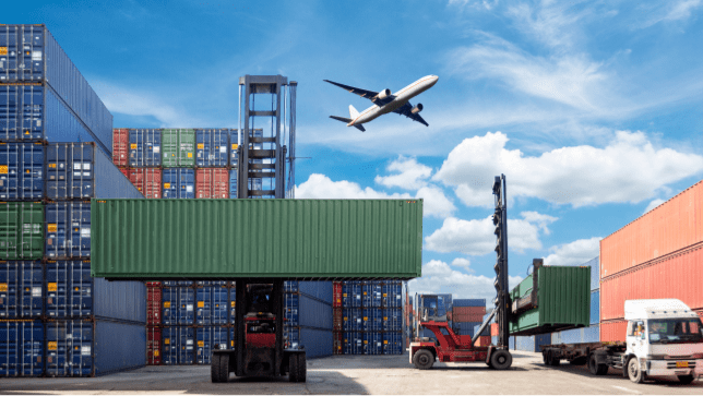 A shipping hard with blue storage crates stacked high as an airplane flies low overhead through a cloud-swept blue sky